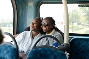 Two women sit smiling on a bus seat, embracing. One wears sunglasses. Sunlight filters through the window, creating a warm, cheerful atmosphere.