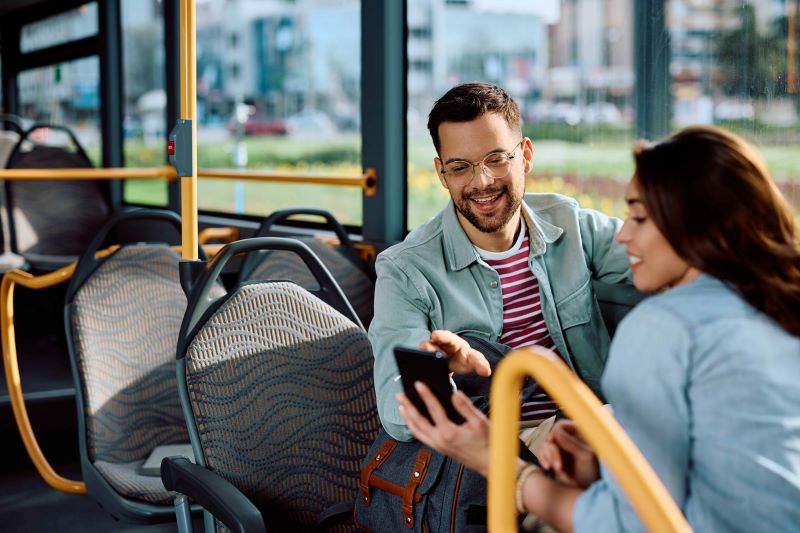 Young man and woman smiling and looking at a smartphone on a sunny bus. The bus interior is bright with yellow railings and patterned seats. Casual and cheerful vibe.