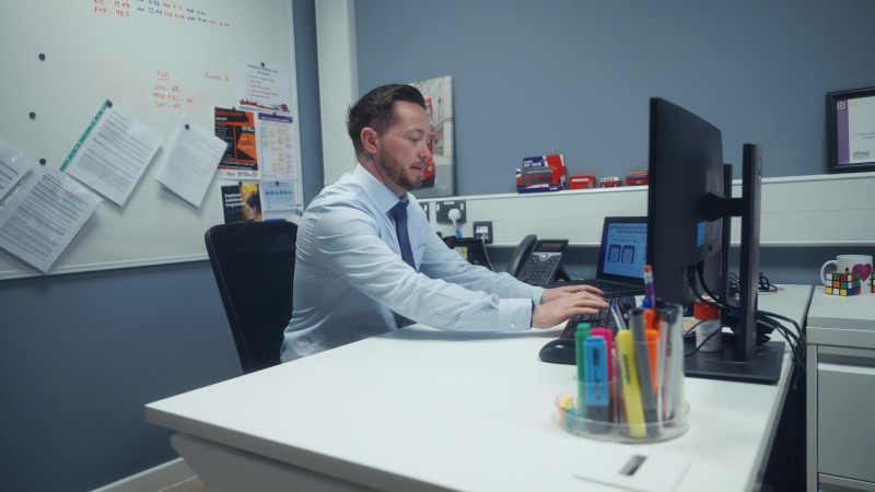 A man in a blue shirt works at a computer in an office. The desk is cluttered with stationery. A whiteboard with papers is in the background. The atmosphere is focused and professional.