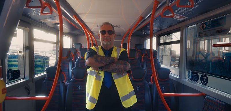A confident man in a safety vest stands with arms crossed inside an empty bus. The brightly lit interior features red poles and patterned seats.