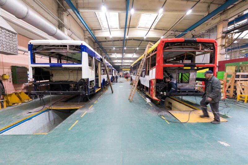 Interior of a bus factory showing two buses under construction in a spacious, well-lit garage. Workers are around, creating a focused, industrious atmosphere.