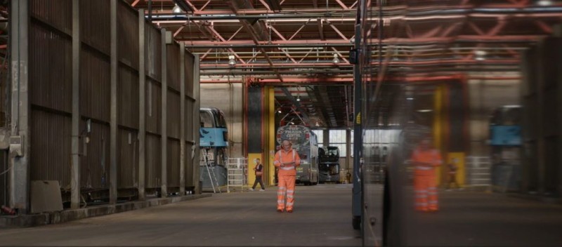 A worker in an orange safety suit stands in a vast train depot, surrounded by parked rail cars and industrial equipment, conveying a sense of industriousness.